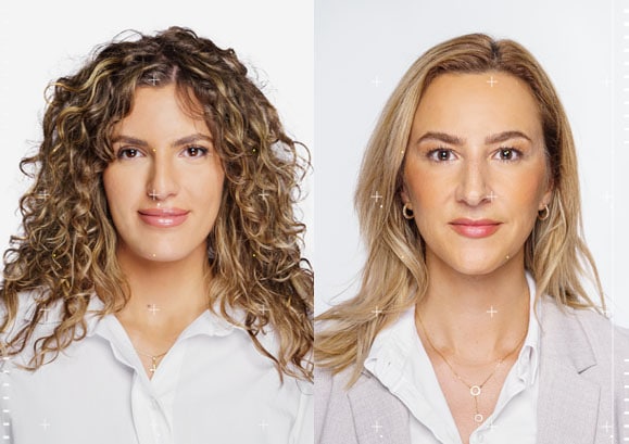 image of two women: Woman on the left is soft smiling with long curly brown hair wearing a white button up shirt. Woman on the right is soft smiling with medium length blonde hair. She is wearing a blazer with a white button up shirt and earrings. 