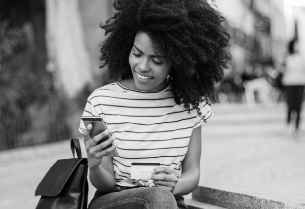 woman with curly hair sitting on sidewalk holding a credit card and smart phone.
