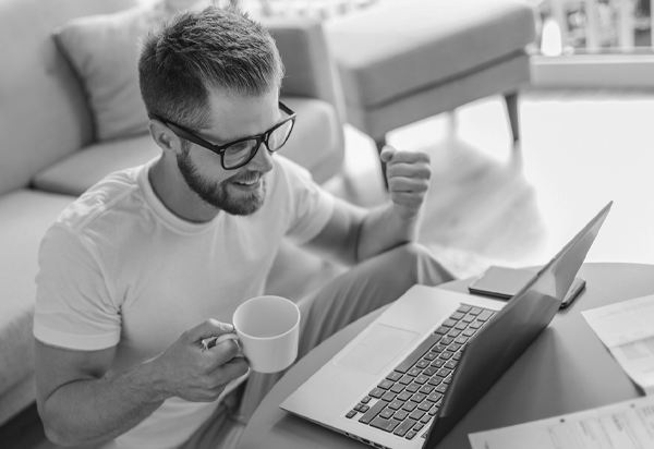 image of man wearing glasses holding a coffee cup in one hand and making a fist in the other hand. Laptop on the table.