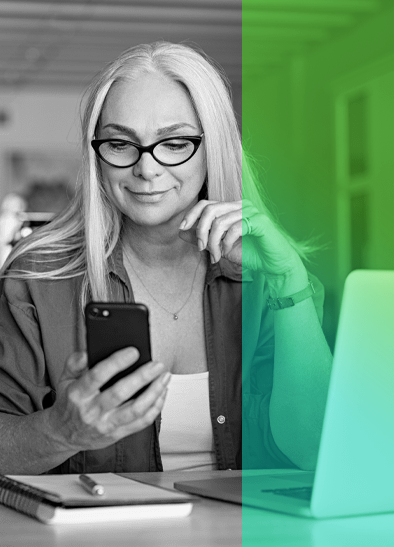 image of woman wearing glasses sitting down at table with computer and on her phone.