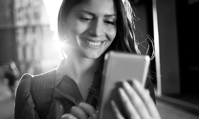 image of woman holding smart device smiling at screen. Photo is in black and white.