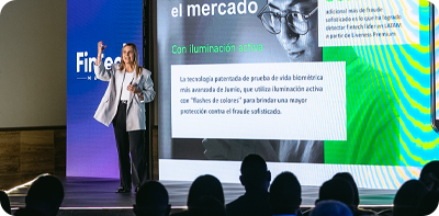 Image of woman on stage presenting to audience. The presentation in the background is showing Jumio content.